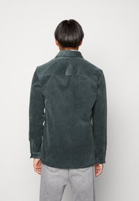 Persona vista da dietro che indossa una giacca di velluto a coste verde scuro e jeans grigi chiari su uno sfondo bianco uniforme.