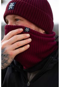 Bordeauxfarbene, gerippte Strickmütze und passendes Nackenband mit Logostick. Merkmale einer eng gestrickten Textur. Schwarze Jacke mit sichtbarem Reißverschluss.