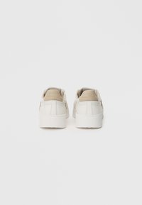 Białe sneakersy z gładkiej skórzanej cholewki, beżowymi zamszowymi akcentami na pięcie oraz teksturowaną gumową podeszwą. Klasyczny niski krój.