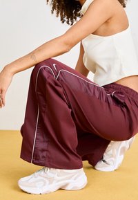 Bordové sportovní kalhoty s bílými bočními pruhy, vyrobené z hladkého materiálu, v kombinaci s masivními béžovými teniskami na žluté podlaze.