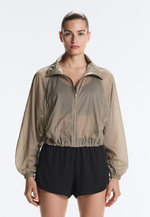 Femme aux cheveux tirés en arrière, portant une veste beige transparente zippée et un short noir, se tenant devant un fond blanc.
