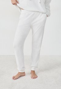 Witte sweatpants met geribbelde boorden en tailleband, met een zachte textuur. Draagt een bijpassende lichtgekleurde top en staat blote voeten op een tapijt.