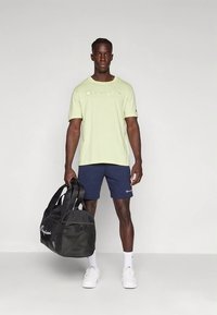Camiseta de algodón verde claro con el logo "Champion", pantalones cortos azul marino, calcetines blancos, y sosteniendo una bolsa de deporte negra con detalles de malla.