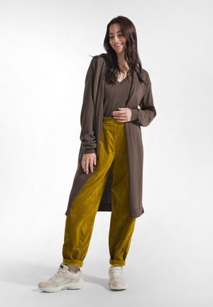 Smilende kvinde iført en lang brun cardigan, brun top, sennepsgule bukser og hvide sneakers står mod en ensfarvet hvid baggrund.