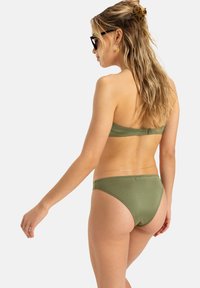 Žalias bikini rinkinys, kuriame yra bandeau stiliaus viršus ir žemas apatinės dalies kirpimas. Lygus paviršius su minimaliais metaliniais detalėmis. Modelis pateiktas iš nugaros.