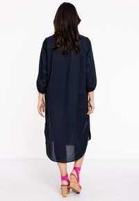 Marineblauwe jurk tot de knie met ¾ mouwen, zijsplitten en een relaxte pasvorm. G gedragen met roze sandaaltjes met sleehak en kurken hakken.