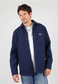 Veste en tissu doux de couleur bleu marine avec boutonnières, dotée d'une poche poitrine, d'une coupe décontractée et de boutons-pression visibles sur le devant.