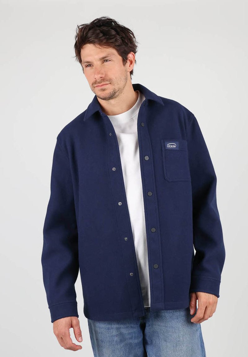 Veste en tissu doux de couleur bleu marine avec boutonnières, dotée d'une poche poitrine, d'une coupe décontractée et de boutons-pression visibles sur le devant.