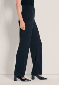 Vrouw in hoge, wijde zwarte broeken met een subtiel gemêleerd patroon en zwarte lakleren block heel schoenen.