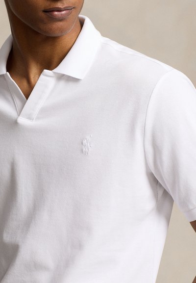 Detailný záber osoby oblečenej v bielej polokošeli z textúrovaného materiálu s malým vyšívaným logom na hrudi.