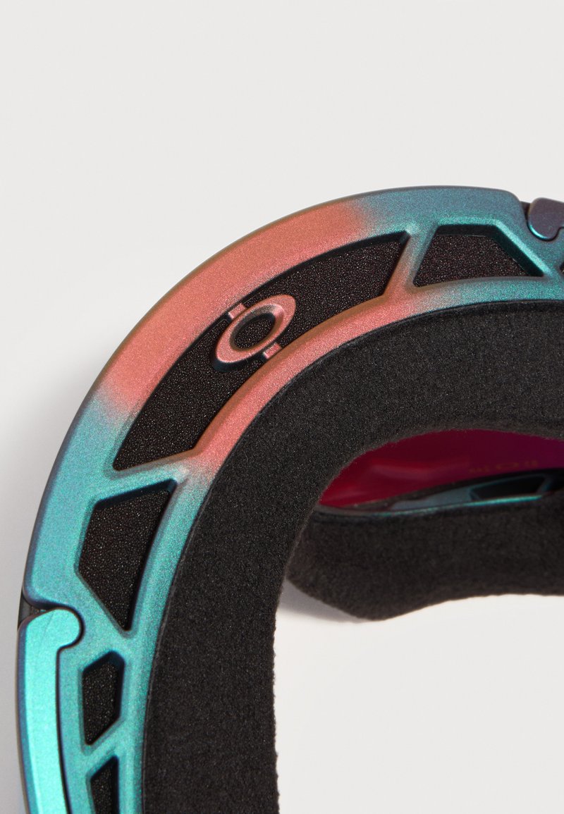 Primer plano del borde del marco de gafas acolchado con espuma iridiscente en azul y rosa, con cortes de ventilación y un pequeño detalle circular de metal.