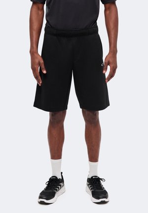 Homme portant un short de sport noir, des chaussettes blanches montantes et des chaussures de course noires et blanches, debout, les bras détendus le long du corps.