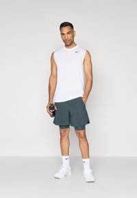 Biela bezrukávová športová košeľa, tmavozelené šortky a biele tenisky. Košeľa má čierne logo Nike. Drží fľašu s vodou.