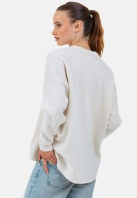 Hellbeiger, oversize Pullover mit strukturierter Webung, überschnittenen Schultern und abgerundetem Saum. Kombiniert mit hellblauen Denim-Jeans.