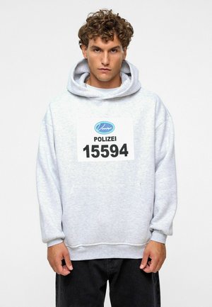 Grijze hoodie van zacht materiaal, met een grote voorzak en een opvallende opdruk "POLIZEI 15594". Zwarte broek.