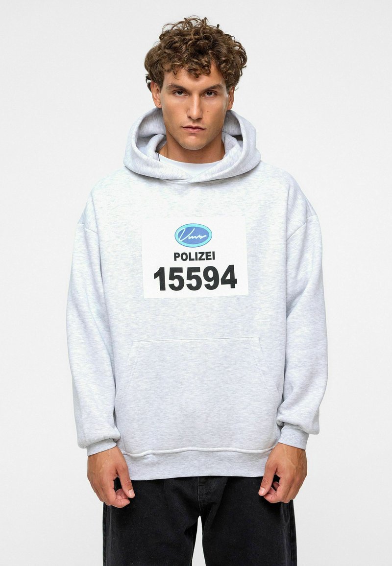Grijze hoodie van zacht materiaal, met een grote voorzak en een opvallende opdruk "POLIZEI 15594". Zwarte broek.