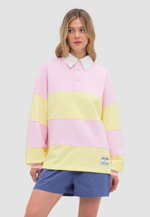 Joven mujer con una camisa polo de manga larga a rayas en tonos rosa pastel y amarillo, y pantalones cortos azules a cuadros, de pie frente a un fondo liso.