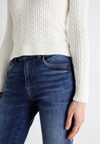 Balta adītu džemperis ar ribotu apakšmalu apvienots ar ziliem džinsa biksēm, kurām ir viegla nodiluma pazīme un kontrastējoša šuvju apdare kabatās.