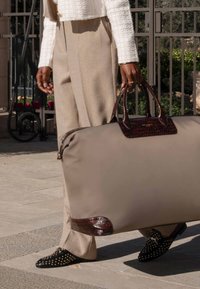 Grand sac de voyage gris avec des poignées et des accents en cuir brun texturé. Porté avec un pantalon taillé de couleur claire et des chaussures noires cloutées.