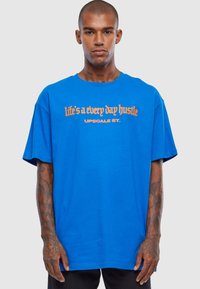 Royal blue oversized t-shirt lavet af bomuld, med orange tekst der står "livet er en daglig hård kamp" og "UPSCale ST." foran.