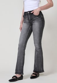 Jeans de denim gris con piernas acampanadas, que presentan bolsillos frontales, un cierre de botón y detalles de tachuelas plateadas en los bolsillos. Textura suave.