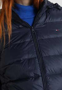 Tmavě modrá péřová bunda se zipem, s vodorovným prošíváním, nastavitelnou kapucí a malým logem na hrudi. Hladká textura.