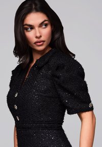 Robe noire texturée avec des manches bouffantes, des sequins et des accents de boutons dorés. Elle présente un col et un design à taille cintrée.