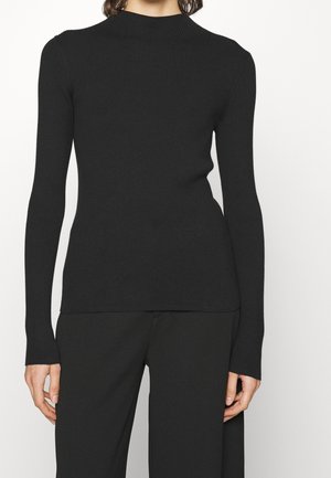 Kvinde iført en tætsiddende sort ribstrikket langærmet top med høj hals, kombineret med sorte bukser mod en ensfarvet baggrund.