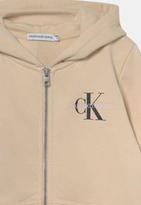 Sudadera con cremallera color beige, fabricada en una suave mezcla de algodón. Presenta un logo bordado en negro de 'CK' y el texto "CALVIN KLEIN JEANS" en el frente.