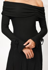 Vestito nero a spalle scoperte in maglia a costine, con maniche lunghe, dettagli arricciati sul colletto e polsini svasati con lacci.