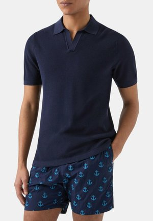 Uomo che indossa una polo a maniche corte testurizzata blu navy e pantaloncini da nuoto blu navy con motivi di ancore azzurri, una mano in tasca.