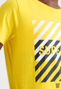 Camiseta de algodón amarilla con un diseño gráfico que presenta franjas diagonales en blanco y negro y el texto "SUPER" en amarillo negrita.