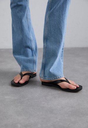 Črne japonke z širokim V-oblikovanim trakom, narejene iz gume. Nosijo se z svetlo modrimi ravnimi kavbojkami, ki prikazujejo teksturiran denim.