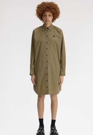 Kvinde med krøllet hår iført en langærmet, knappet kjole med små tern og sorte sokker med sorte loafers, stående oprejst.