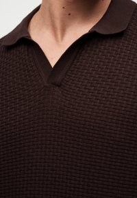 Κοντινή λήψη ενός ατόμου που φοράει καφέ σκουρόχρωμο πλεκτό πόλο μπλουζάκι με μαλακό γιακά και άνοιγμα σε σχήμα V.