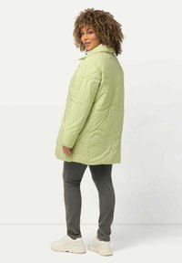 Abrigo acolchado verde con patrones de corazones, cuello alto y diseño de longitud media. Combinado con jeans grises y zapatillas blancas.