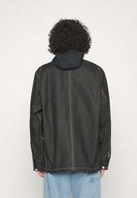 Chaqueta de denim negro con capucha, costuras blancas y puños a presión. Presenta un ajuste holgado y un diseño de espalda recta.