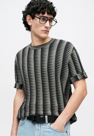 Pull à manches courtes rayé gris et noir avec une coupe ample, un col côtelé et un tissu en maille doux et texturé. Porté avec un denim clair et une ceinture noire.