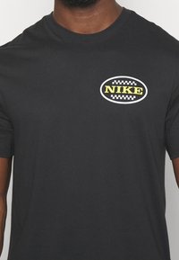 T-shirt en coton noir avec un logo "NIKE" rond blanc et jaune avec une bordure à carreaux sur la partie gauche de la poitrine. Manches courtes.
