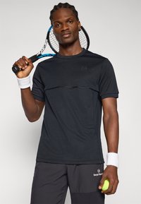 Černé sportovní tričko s krátkým rukávem a jemným designovým detailem přes hruď, spárované se šedými šortkami. Drží tenisovou raketu a míček.