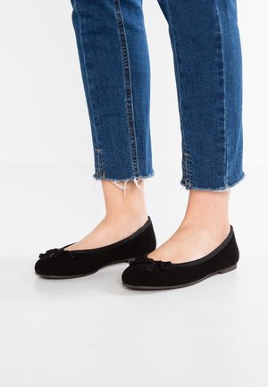 Ben iført blå jeans med opsmøgede kanter og sorte ballerinaer med dekorative sløjfer, stående på en glat hvid gulv.