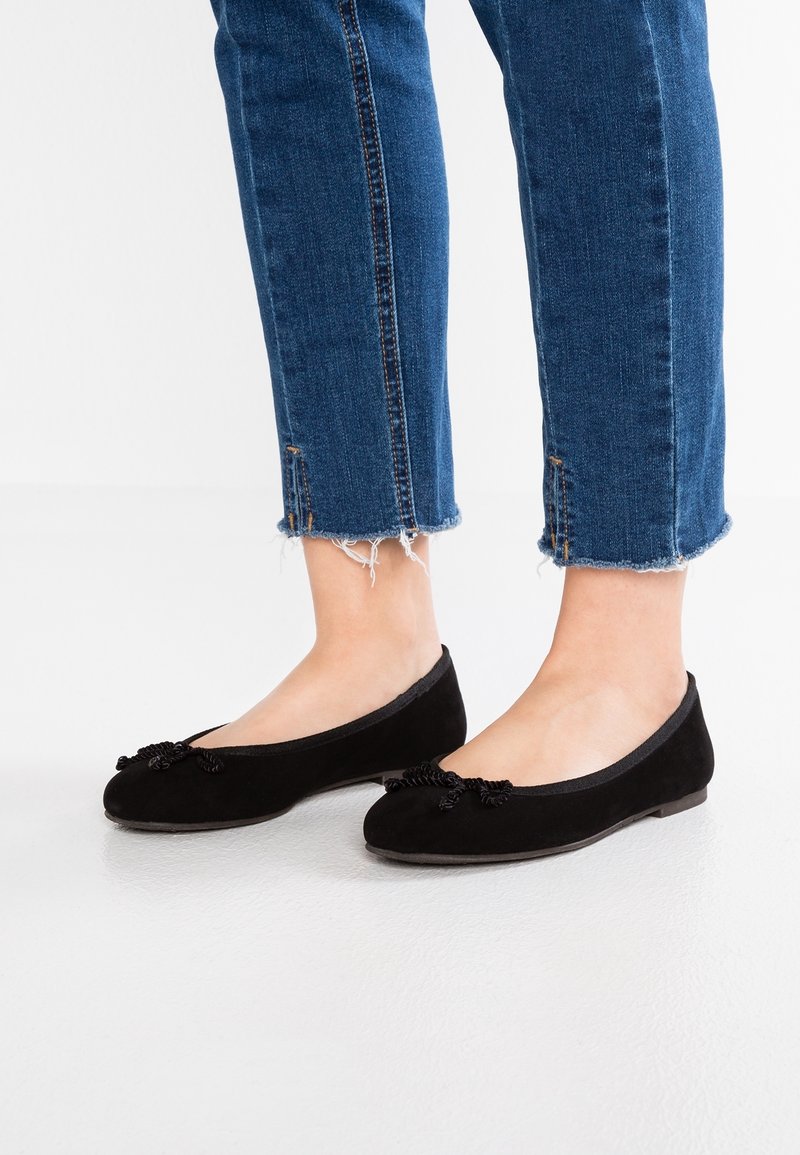 Jambes portant un jean bleu à ourlet effiloché et des ballerines noires ornées de nœuds décoratifs, se tenant sur un sol blanc lisse.