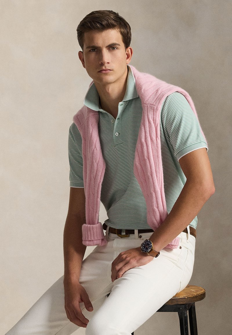 Light pink cable-knit sweater vest over a green and white striped polo, paired with white pants and a brown leather belt, showcasing a watch.