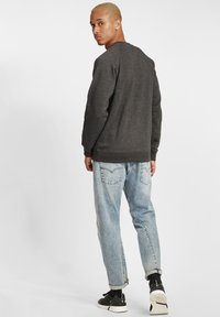 Grijze sweatshirt met een ronde hals en geribde mouwen, gecombineerd met lichtblauwe opgerolde jeans en zwarte sneakers. Achtergrond is wit.