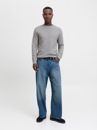 Man standing wearing a light gray textured sweater, wide-leg blue jeans with a black belt, and black slip-on shoes against a white background.