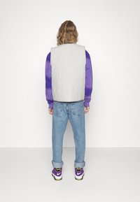 Gepolstertes graues Vest über einem lila Batik-Sweatshirt, kombiniert mit hellblauen Jeans und schwarzen Schuhen mit lila und gelben Akzenten.