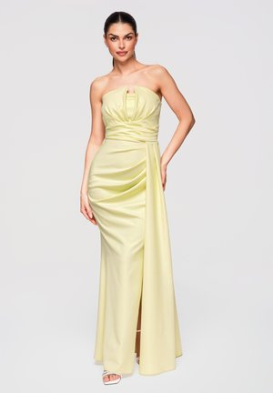 Kvinde poser i en stropløs, lysegul satin kjole med draperede detaljer og en frontslids, iført hvide åbne tå-sandaler.