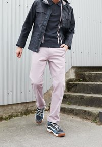 Dark, zippered jacket over a navy sweater, paired with light pink pants and blue sneakers. Grey background with metal siding.