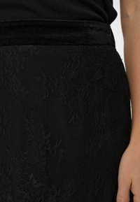 Melna svārku ar mežģīņu pārklājumu, kas rotāta ar teksturētu dizainu un samta jostu. Tuvplānā redzami smalki ziedu raksti uz sieta fona.