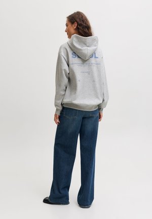 Femme debout, de dos, portant un sweat à capuche gris avec le texte "SEOUL" et un jean bleu à jambes larges sur un fond clair uni.